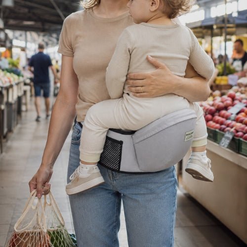 Tabouret porte bebe gris clair avec ceinture réglable, enfant face au porteur dans un marché