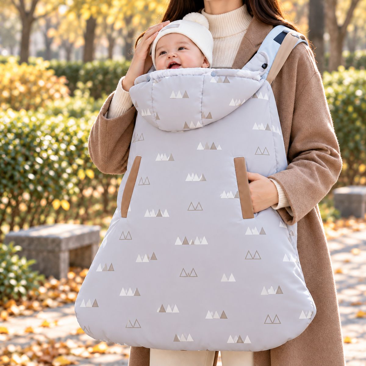 Cape porte-bébé gris clair à motifs montagnes, avec capuche intégrée et ouvertures latérales pour les mains, portée en position ventrale avec bébé.