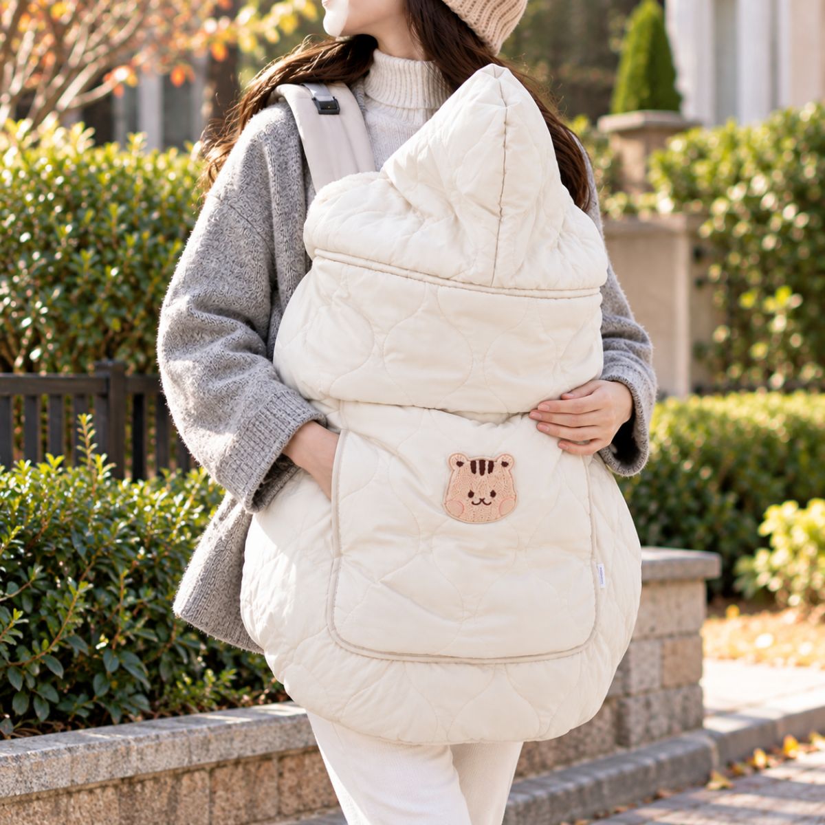 Cape porte-bébé hiver beige à capuche, portée devant une femme, avec tissu matelassé et motif écureuil sur la poche avant.