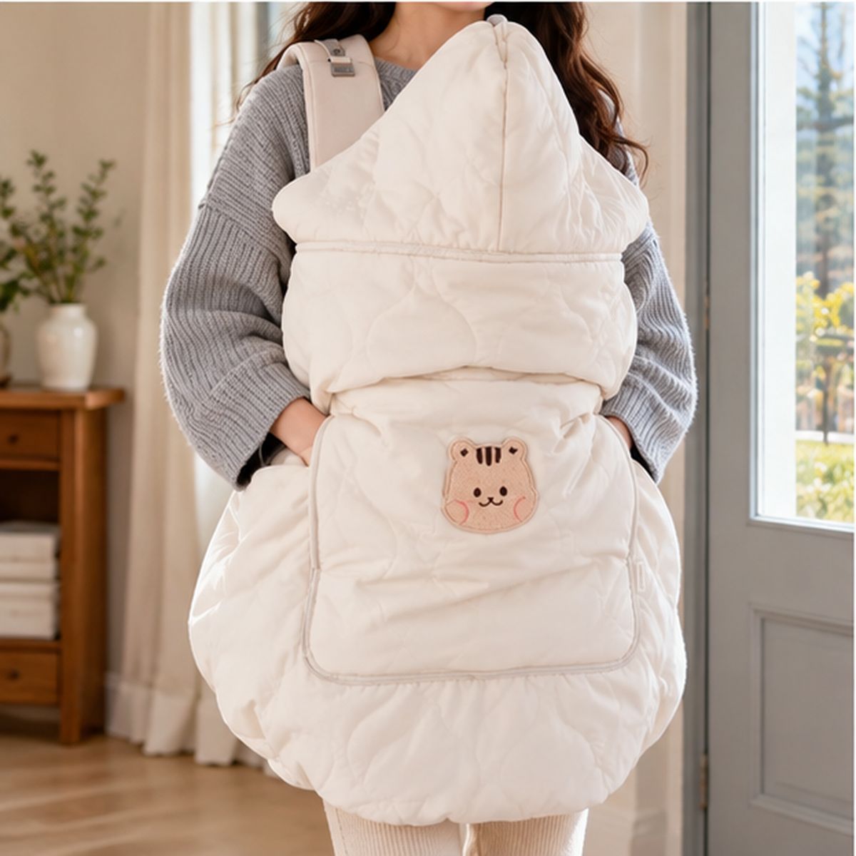Cape porte-bébé hiver beige vue de face en intérieur, avec capuche relevée, matelassage et motif écureuil brodé sur la poche.