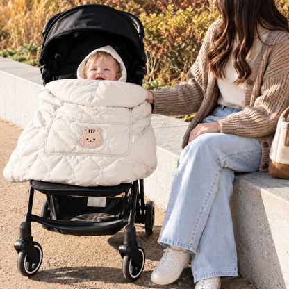 Cape porte-bébé hiver beige utilisée sur une poussette, avec bébé au visage visible, tissu matelassé et motif écureuil sur la poche.