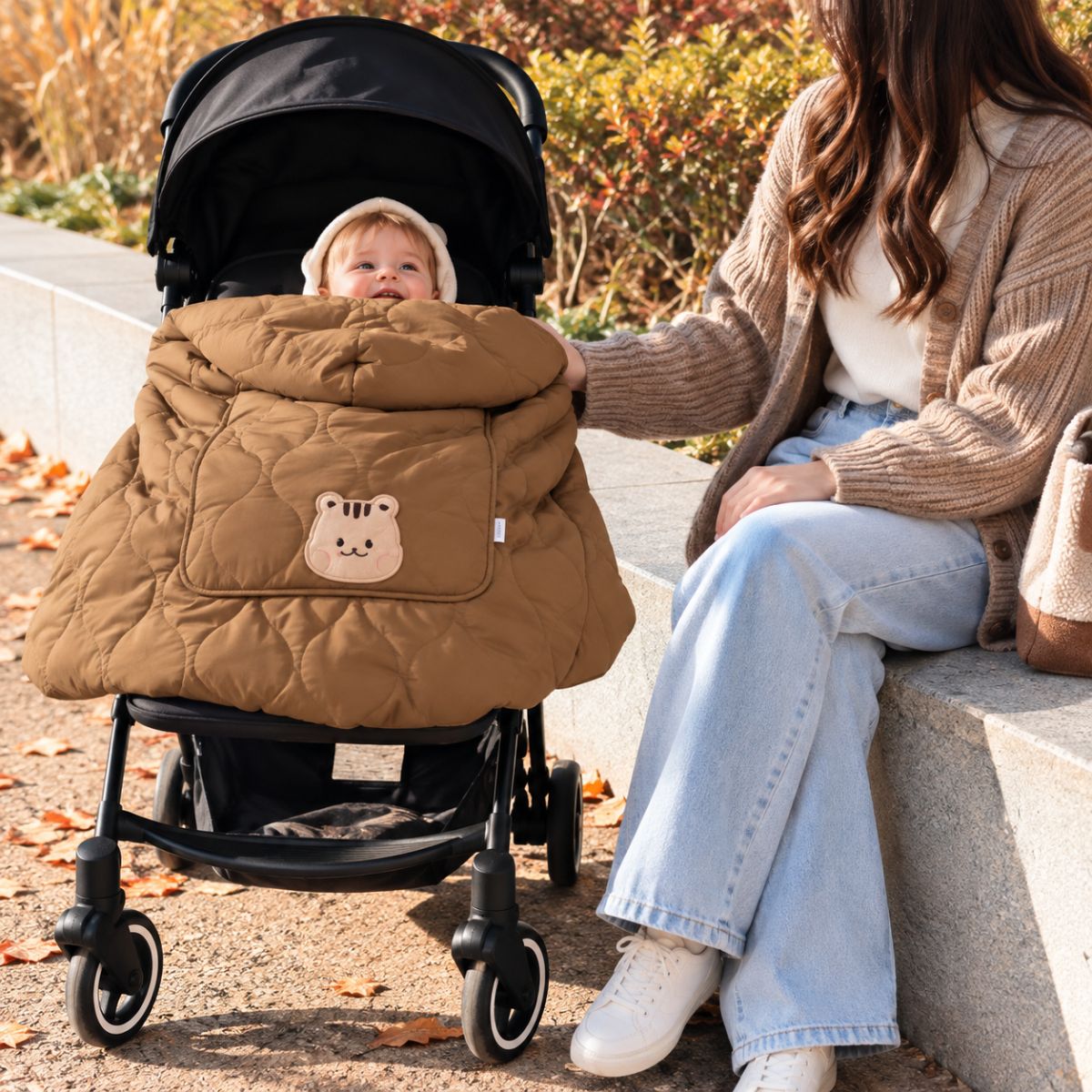 Cape porte-bébé hiver écureuil café installée sur une poussette, avec bébé au visage visible, tissu matelassé et motif écureuil sur la poche.