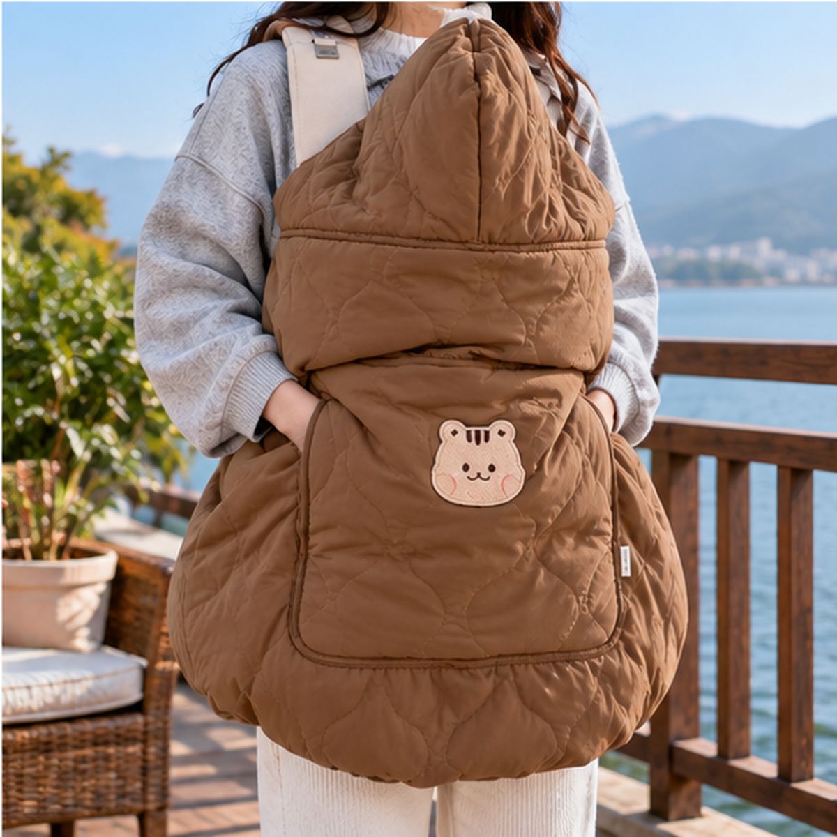 Cape porte-bébé hiver écureuil café, avec capuche relevée, tissu matelassé et poche à motif écureuil, photographiée près d’un lac.