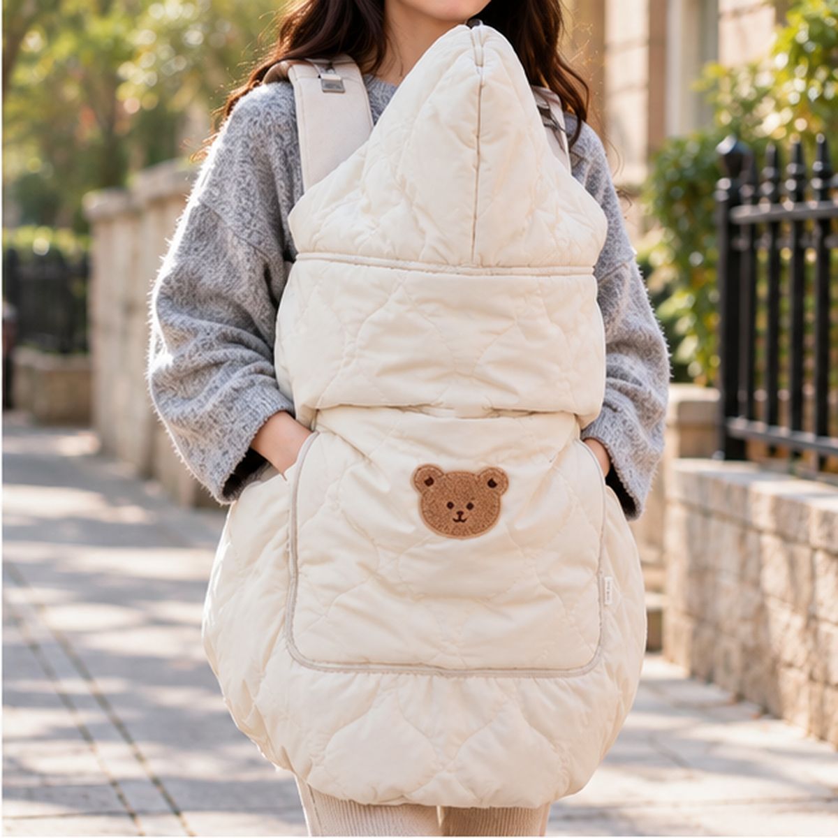 Cape porte-bébé hiver ours beige vue de face en intérieur, avec grande capuche relevée, matelassage et motif ours sur la poche.