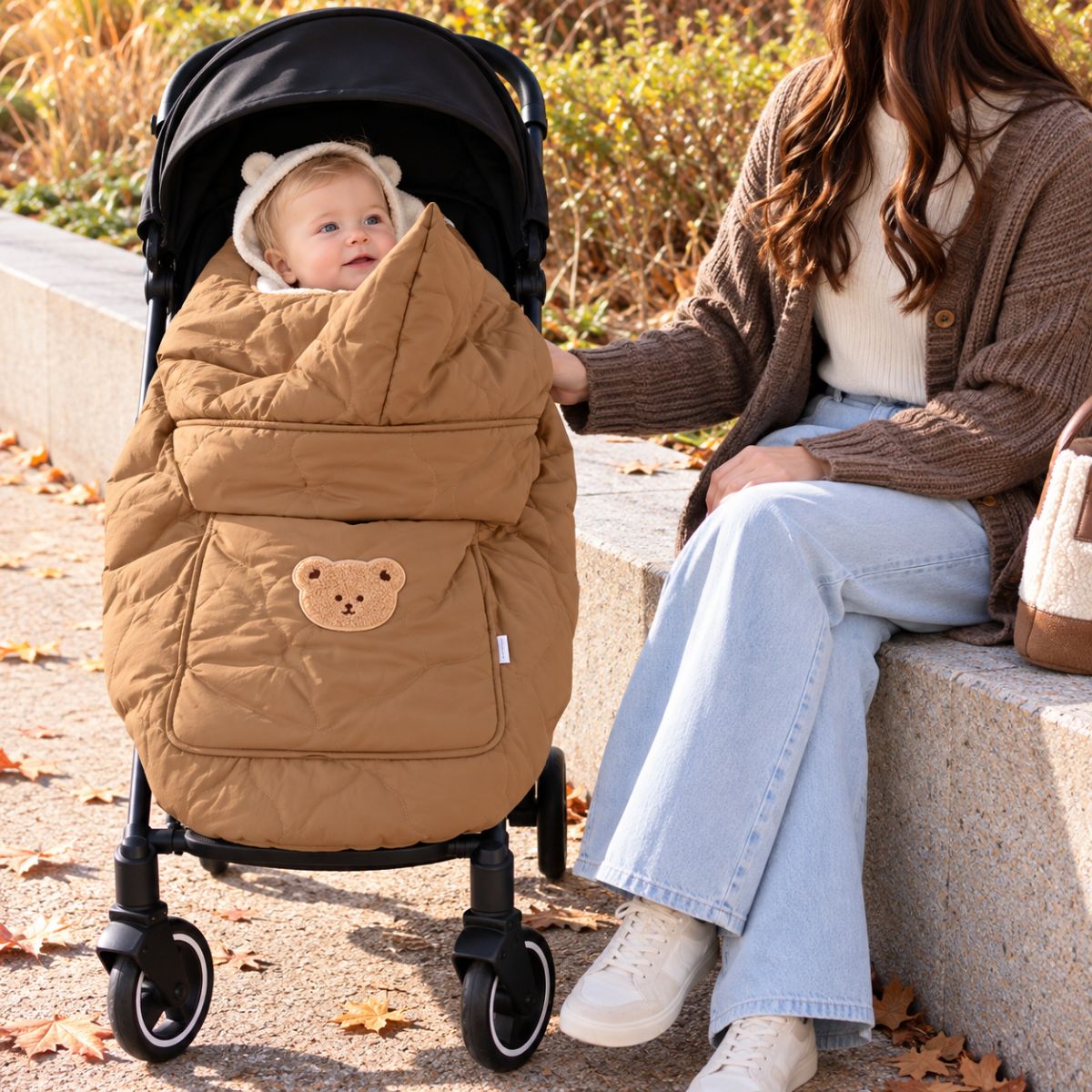 Cape porte-bébé hiver café installée sur une poussette, avec bébé visible, tissu matelassé et motif ours sur la poche avant.