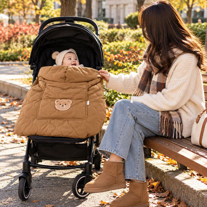 Cape porte-bébé hiver café utilisée sur une poussette en promenade, avec capuche, matelassage et motif ours, près d'une maman assise.