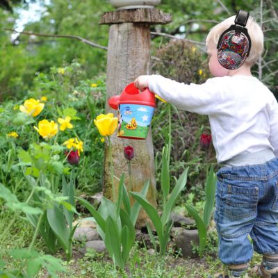 Petit garçon portant un casque anti-bruit enfant noir avec motifs musicaux multicolores SilenceZen, arrosant des fleurs dans le jardin, conçu pour une protection auditive fiable en extérieur