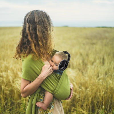 Une mère portant son bébé dans un champ de blé, le bébé équipé d’un casque antibruit enfant noir SilenceAnge, pour un moment de calme et de sérénité en pleine nature