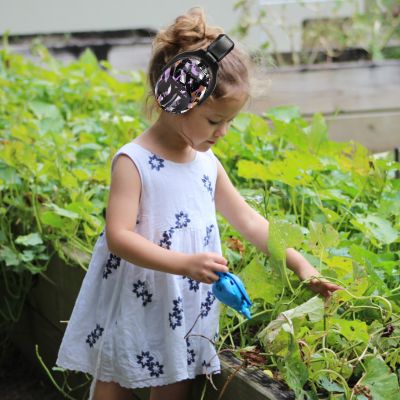 Petite fille portant un casque anti-bruit enfant noir avec motifs violet et orange SilenceZen, jardinant dans la nature, parfait pour protéger ses oreilles tout en s'adaptant à ses besoins