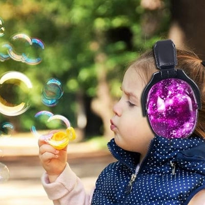 Petite fille soufflant des bulles dans un parc, équipée d’un casque anti bruit enfant violet au design galaxie, parfait pour les activités extérieures et le confort auditif