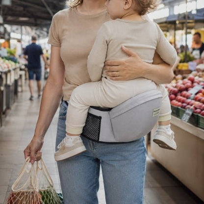 Tabouret porte bebe gris clair avec ceinture réglable, enfant face au porteur dans un marché