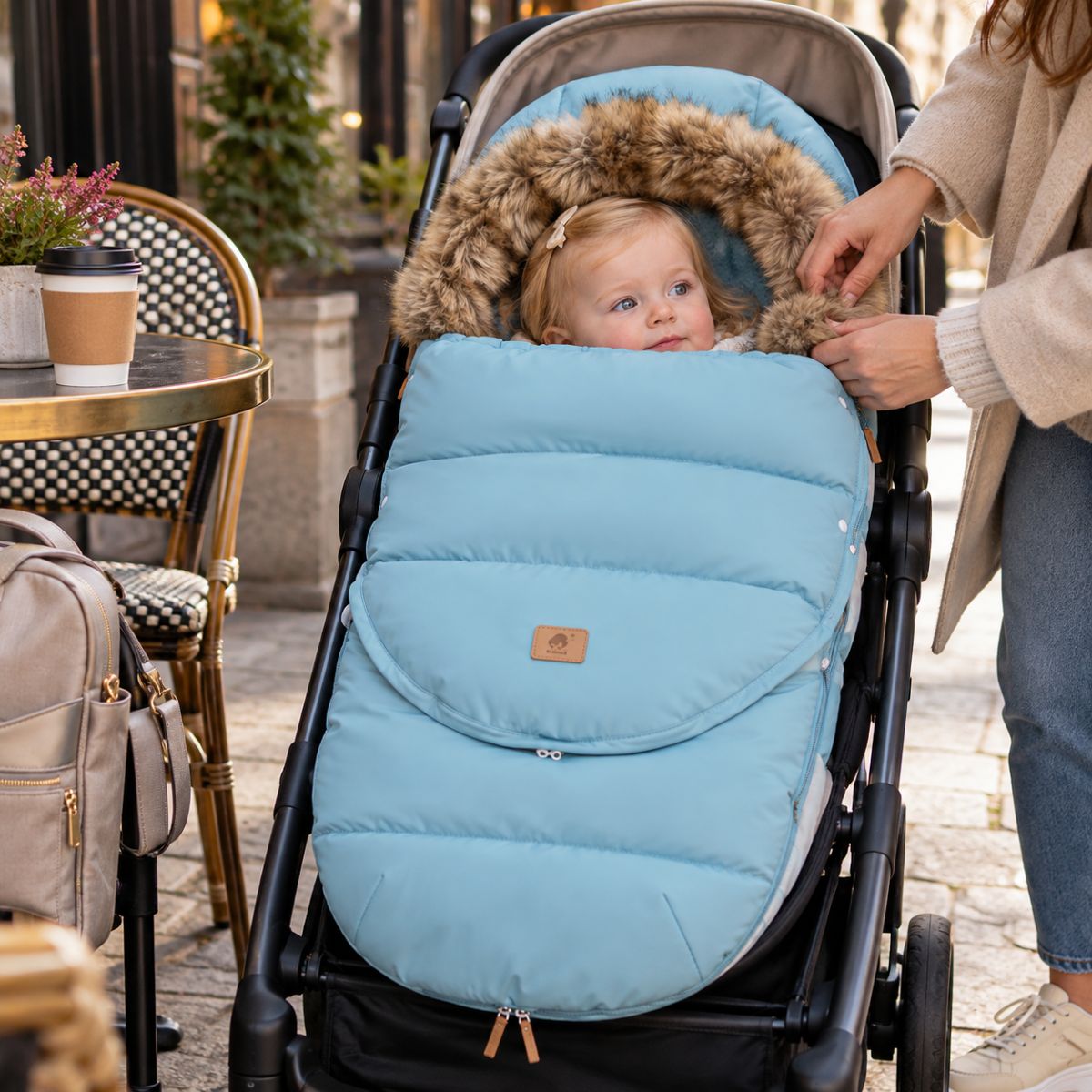 Chancelière poussette bleu clair avec col effet fourrure, bébé installé dans une poussette près d’une terrasse avec sa maman