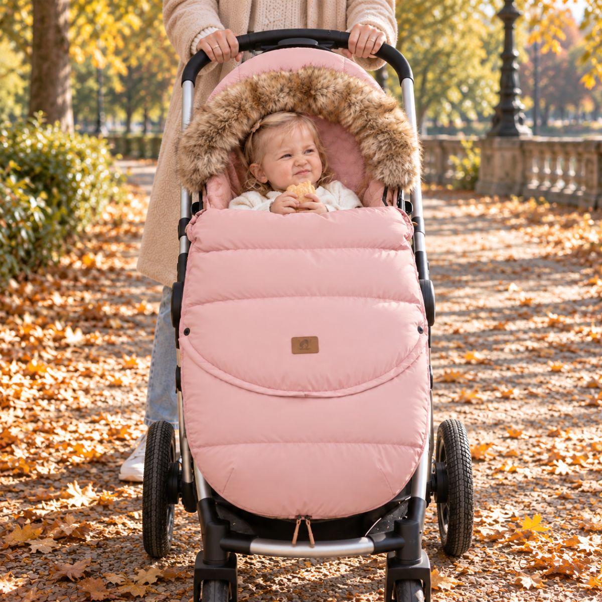 Chancelière poussette rose poudré avec col effet fourrure, bébé installé dans une poussette vue de face dans un parc en automne