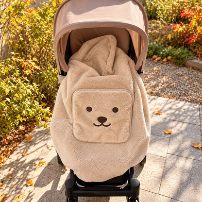 Couverture de portage moumoute beige installée sur une poussette, avec capuche intégrée, tissu pelucheux et grande poche décorée d’un visage d’ours