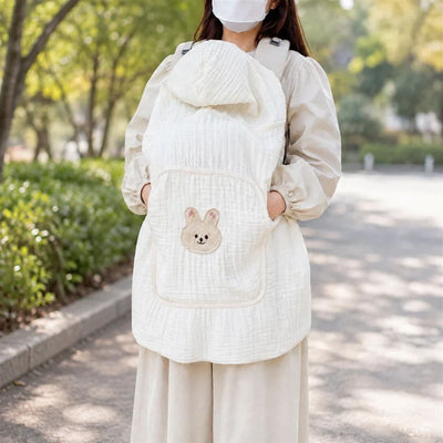 Couverture porte-bébé légère en mousseline écrue avec capuche et poche frontale à motif lapin, portée en extérieur