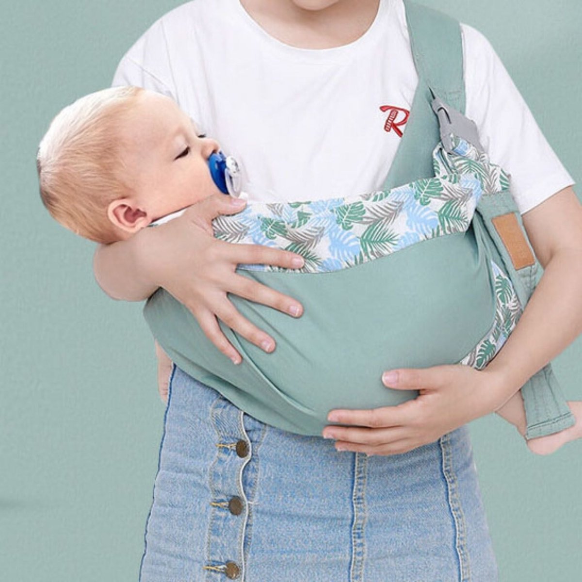 echarpe de portage vert menthe avec bébé blond en position berceau, bord imprimé feuillage bleu vert sur fond uni doux