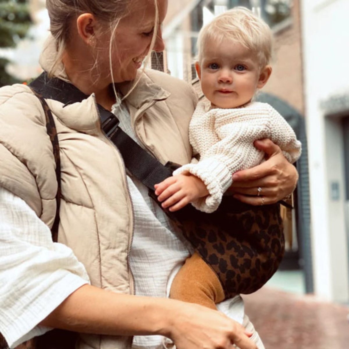 Bébé blond assis dans une écharpe de portage léopard tenue par sa maman