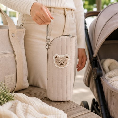 Étui matelassé pour biberon beige avec écusson ourson porté par sa sangle près d’une poussette