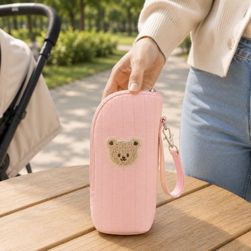 Étui matelassé pour biberon rose avec écusson ourson tenu à la main près d’une poussette