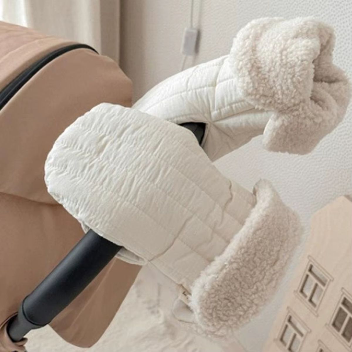 Paire de gants pour poussette beige avec extérieur surpiqué et revers pelucheux, installés sur un guidon noir
