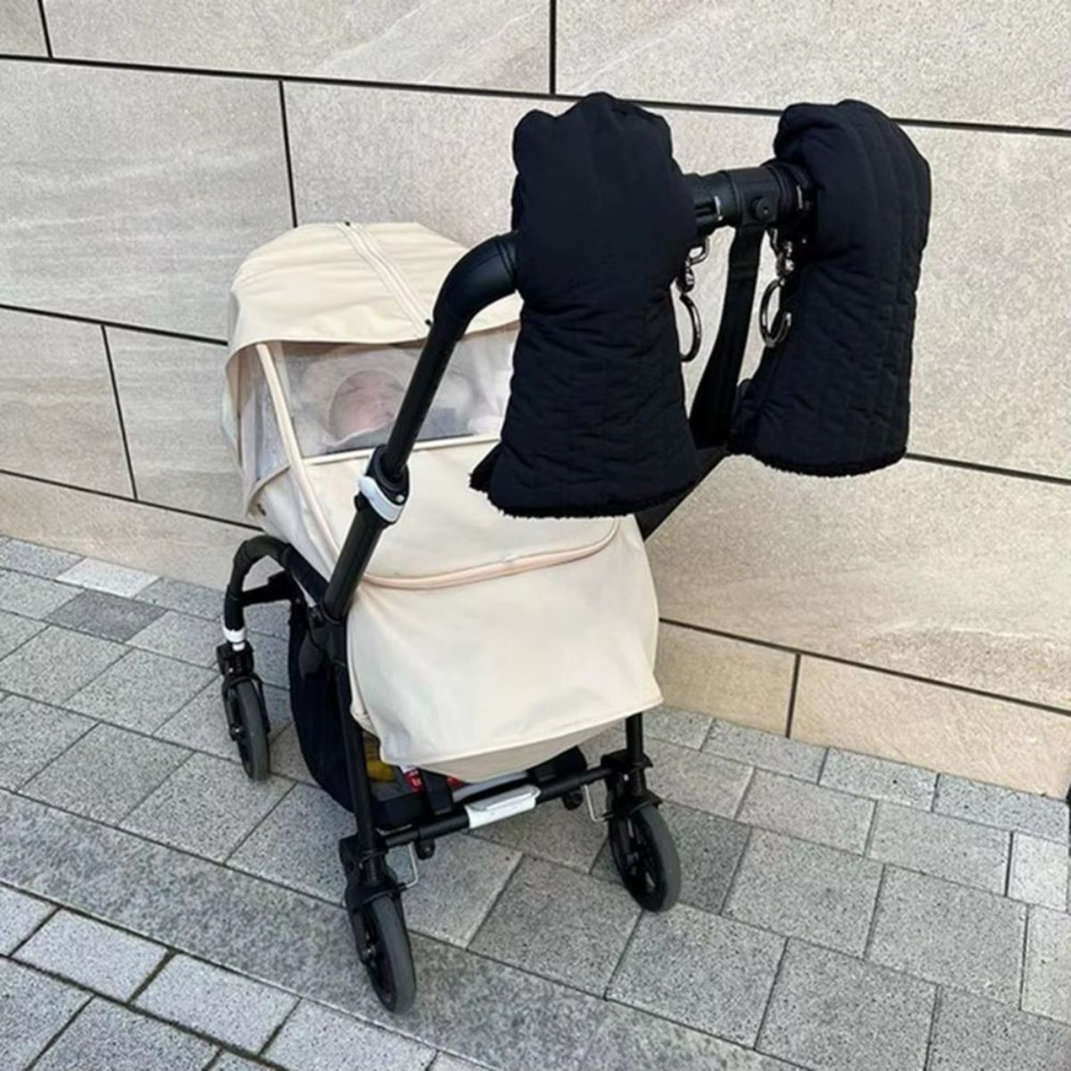 Paire de gants de poussette noirs surpiqués, suspendus au guidon d'une poussette avec habillage beige
