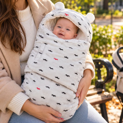Nid d’ange bébé clair à petits motifs noirs, avec capuche arrondie à deux petites oreilles et rabat frontal, tenu dans les bras