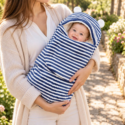Nid d’ange bébé à rayures bleu marine et blanches, avec capuche arrondie à petites oreilles, tenu dans les bras au jardin