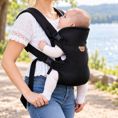 Porte-bébé à partie haute pliable noir avec tête d’ourson brodée beige et boucles latérales, porté près d’un lac