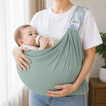 Porte-bébé à une épaule vert sauge avec assise large et sangle réglable, bébé avec biberon en intérieur