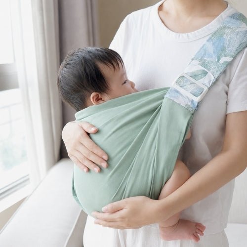 Porte-bébé à une épaule vert sauge avec assise large et sangle réglable, près d’une fenêtre