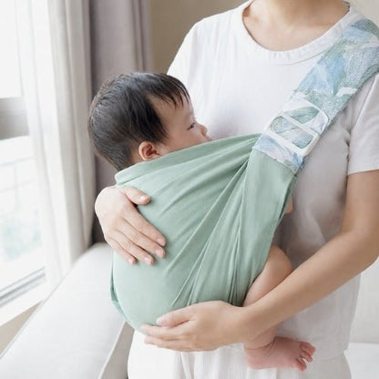 Porte-bébé à une épaule vert sauge avec assise large et sangle réglable, près d’une fenêtre