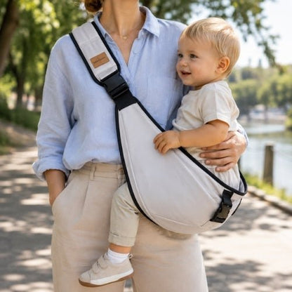 Porte-bébé à une épaule gris clair avec sangle large et réglable, porté latéralement au bord d’un lac