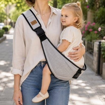 Porte-bébé à une épaule en maille gris clair avec sangle large et réglable, porté latéralement dans une rue