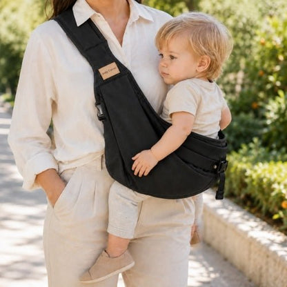 Porte-bébé à une épaule noir avec sangle large et réglable, porté latéralement dans un parc