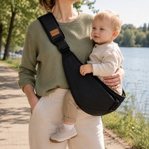 Porte-bébé à une épaule noir avec sangle large et réglable, porté latéralement au bord d’un lac