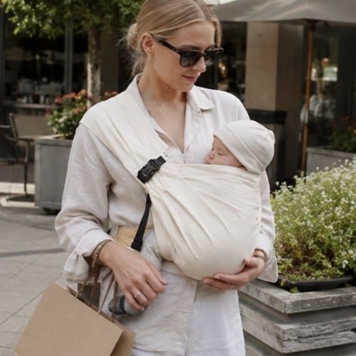 Porte-bébé à une épaule beige avec boucle réglable, utilisé en position frontale près d’un café