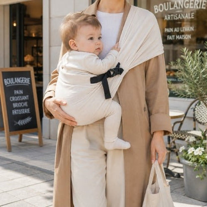 Porte-bébé à une épaule beige avec boucle réglable, port frontal devant une boulangerie