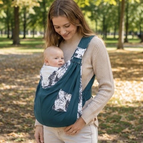 Porte-bébé à une épaule bleu pétrole avec poche intégrée et boucle réglable, porté dans un jardin