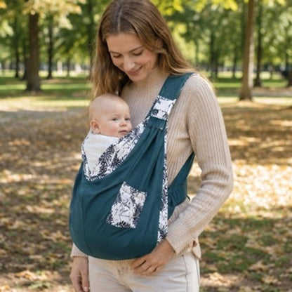 Porte-bébé à une épaule bleu pétrole avec poche intégrée et boucle réglable, porté dans un jardin