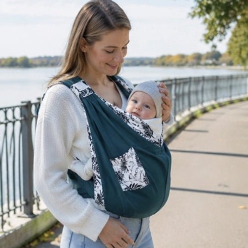 Porte-bébé à une épaule bleu pétrole avec poche intégrée et boucle réglable, porté au bord d’un lac