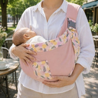Porte-bébé à une épaule rose avec poche intégrée, boucle réglable et bordure florale près d’une terrasse