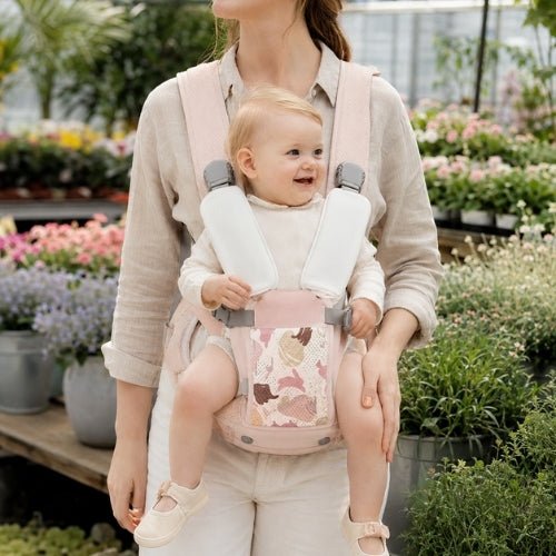 Porte-bébé avec siège détachable rose, porté face vers l’avant près d’un étal de fleurs
