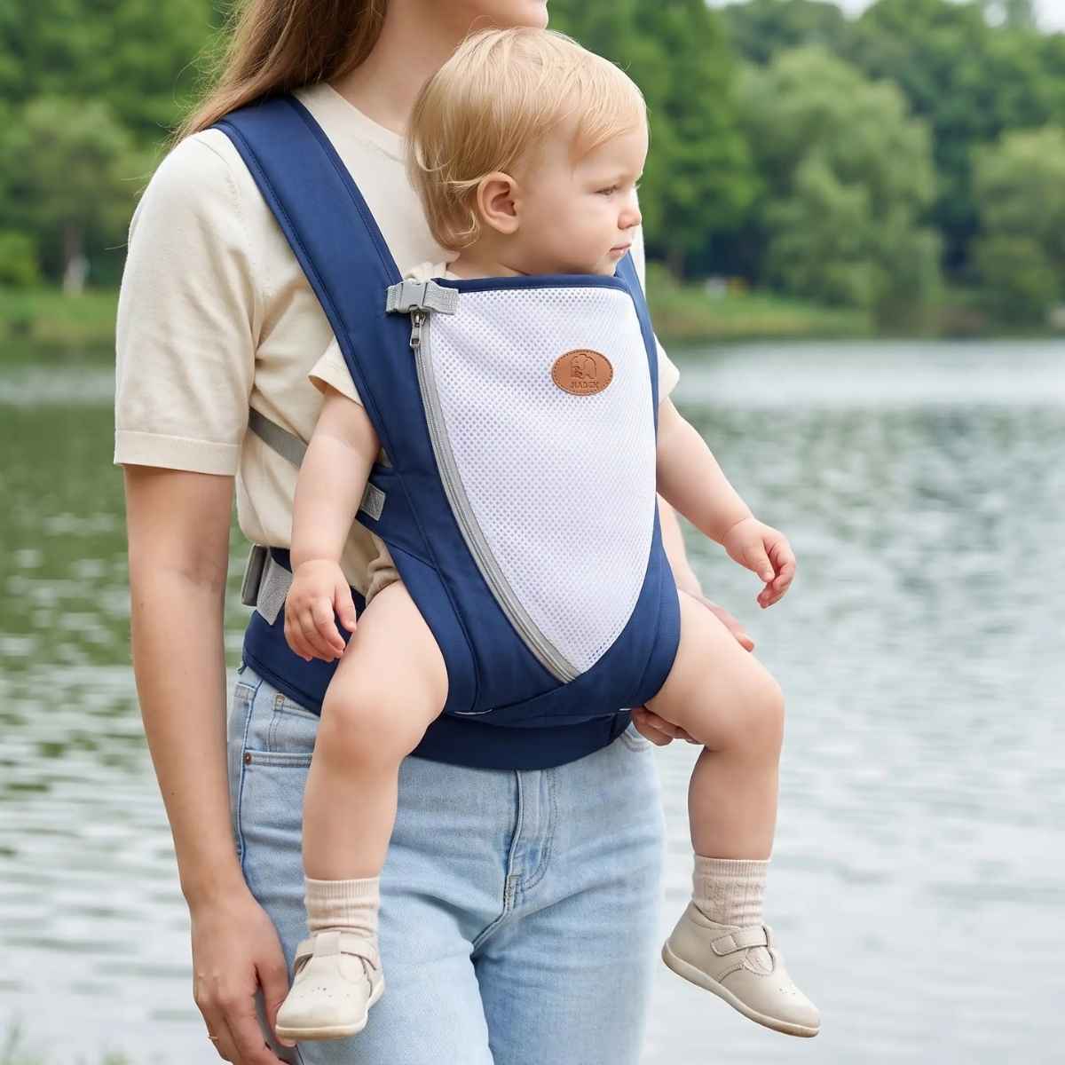 Porte-bébé croisé dans le dos bleu marine avec bretelles larges et panneau en maille, porté face vers l’avant près d’un lac