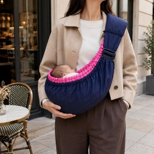 Porte bébé hamac bleu marine avec intérieur rose à carreaux, contour rembourré et sangle réglable près d’un café
