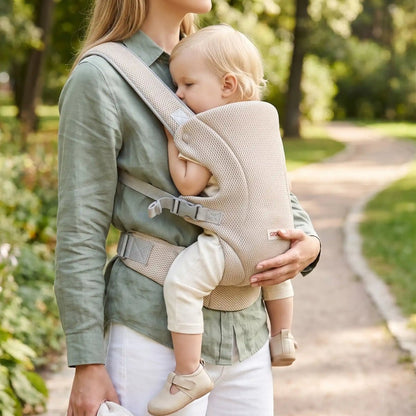 Porte-bébé mesh beige avec bretelles croisées et format léger, porté face au porteur dans un parc