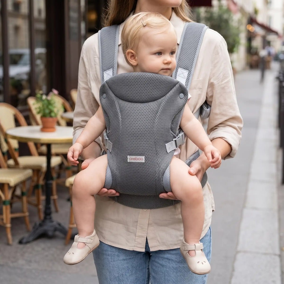 Porte-bébé mesh gris avec bretelles croisées et format léger, porté face vers l’avant près d’un café