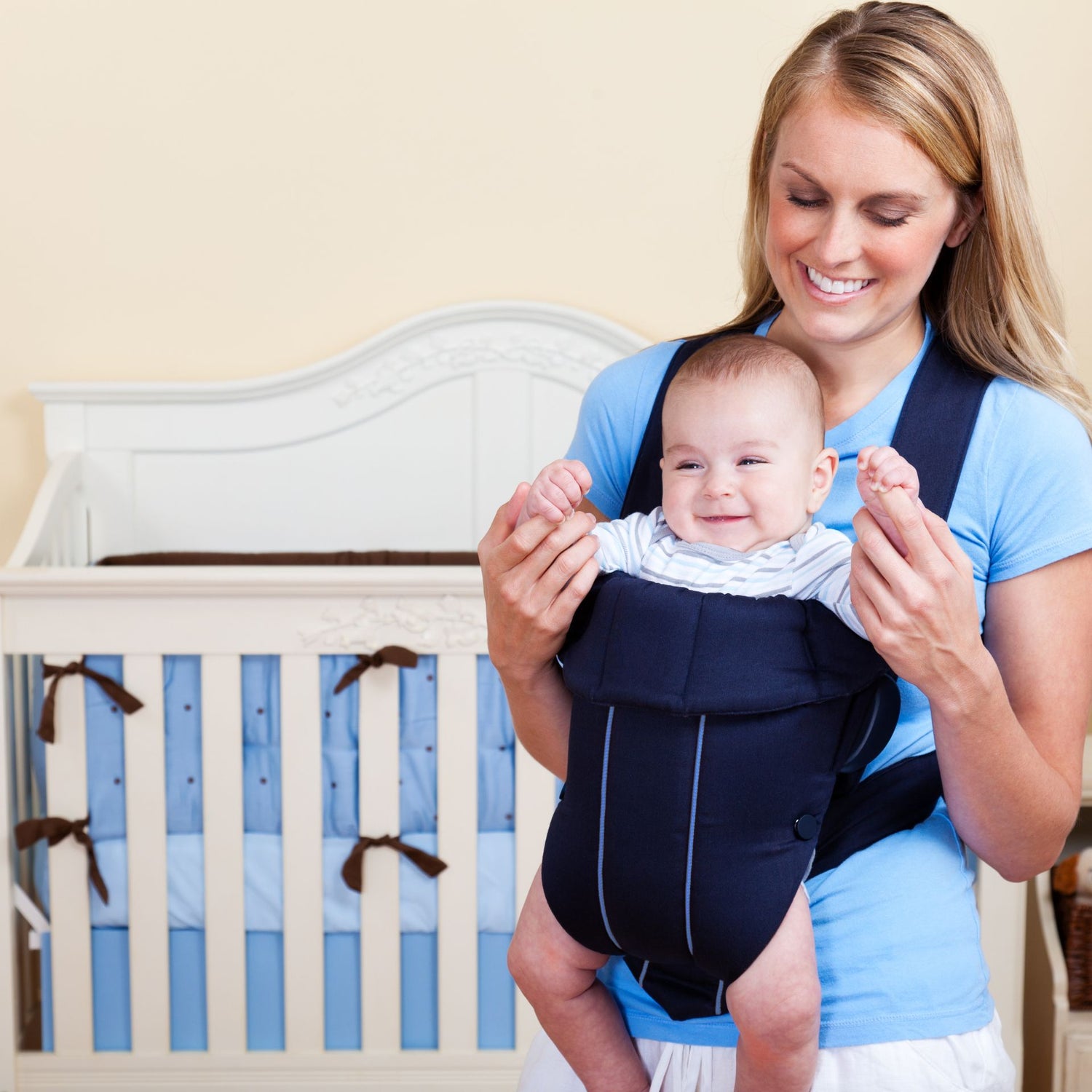 Une maman souriante porte son bébé dans un porte bébé ventral bleu marine, devant un lit de bébé – confort et proximité signés MamanPépite.