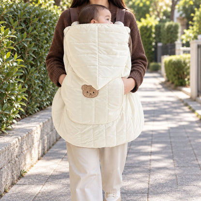 Housse porte-bébé blanc crème matelassée, portée devant une maman en extérieur, avec capuche intégrée, écusson ours et bébé au visage visible