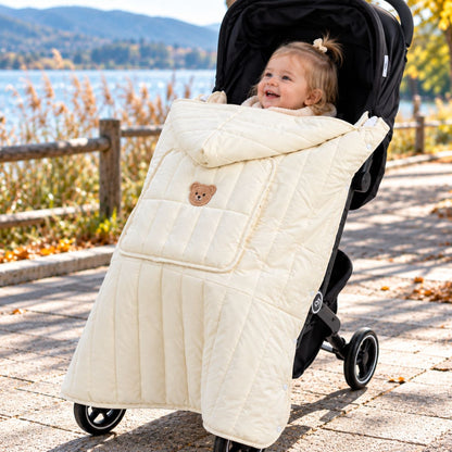 Protège porte-bébé doudoune blanc crème installé sur une poussette, avec grande poche avant, boutons-pression visibles et bébé souriant