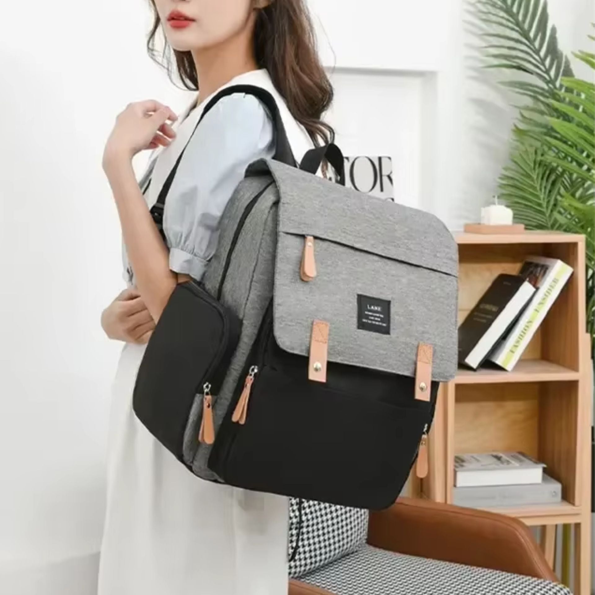 Femme portant un sac à dos à langer gris et noir avec poches latérales et finitions en cuir, dans un intérieur moderne